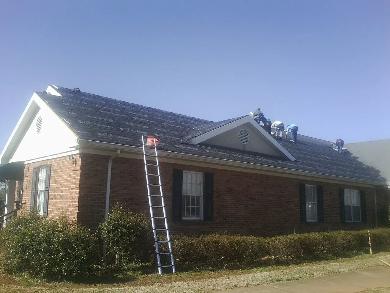 Crew tearing shingles off a brick ranch roof with a ladder set — Emergency Roof Repair work by our crew in Erick