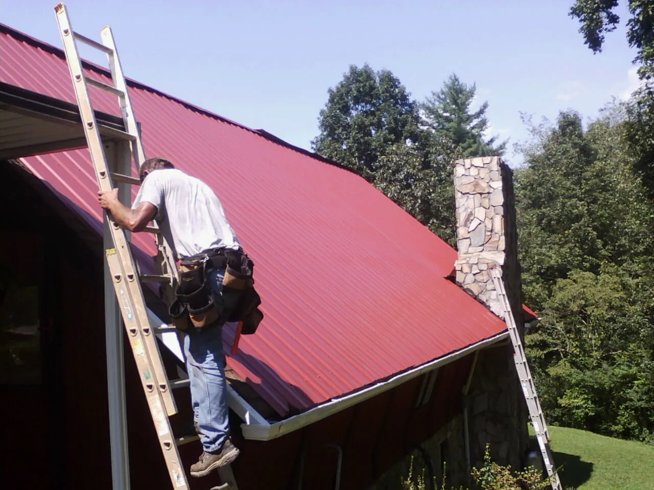 Roofer on a ladder beside a red metal roof and stone chimney — Soffit & Fascia Repair by Roofing Brothers in Erick
